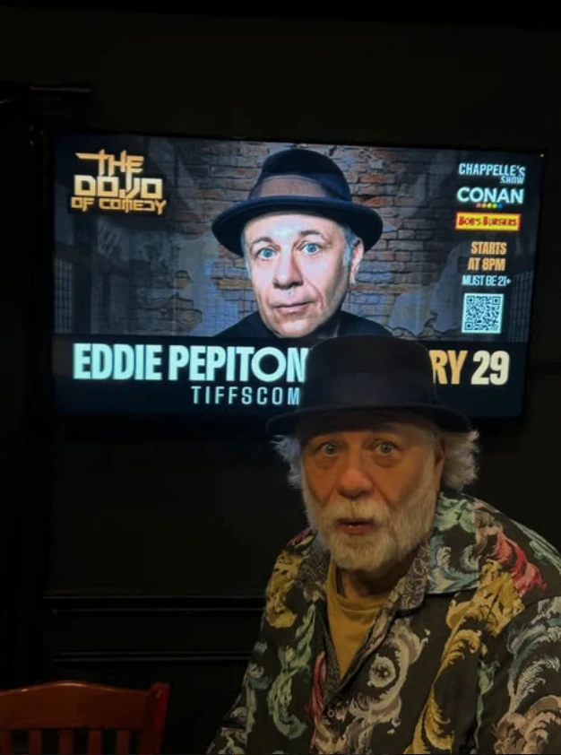 Comedian Eddie Pepitone standing in front of a Dojo of Comedy flyer designed by Celestial Productions